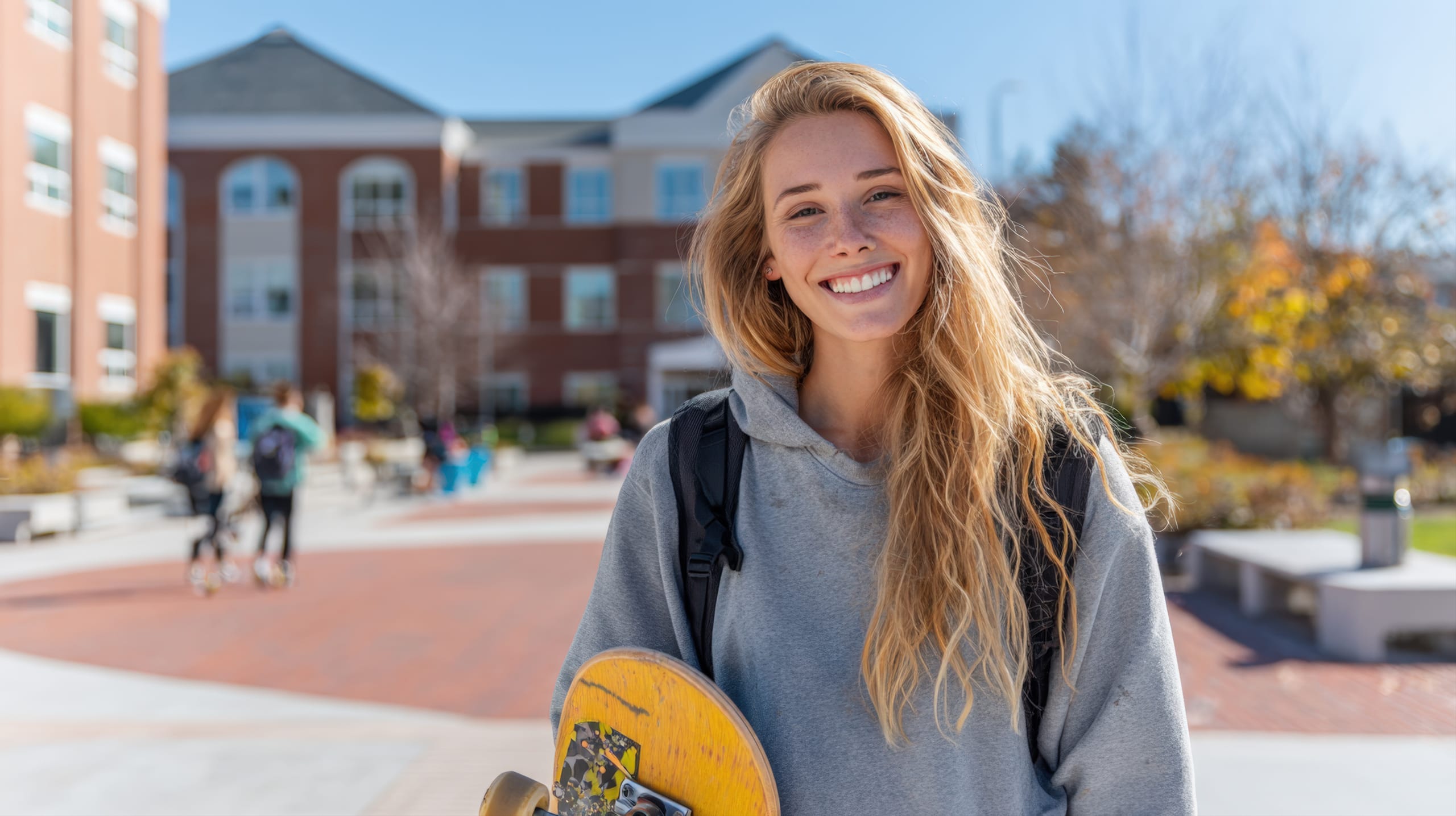 Eine fröhliche Studentin mit langen, welligen Haaren und einem Rucksack lächelt strahlend, während sie ein Skateboard auf einem Universitätscampus hält.